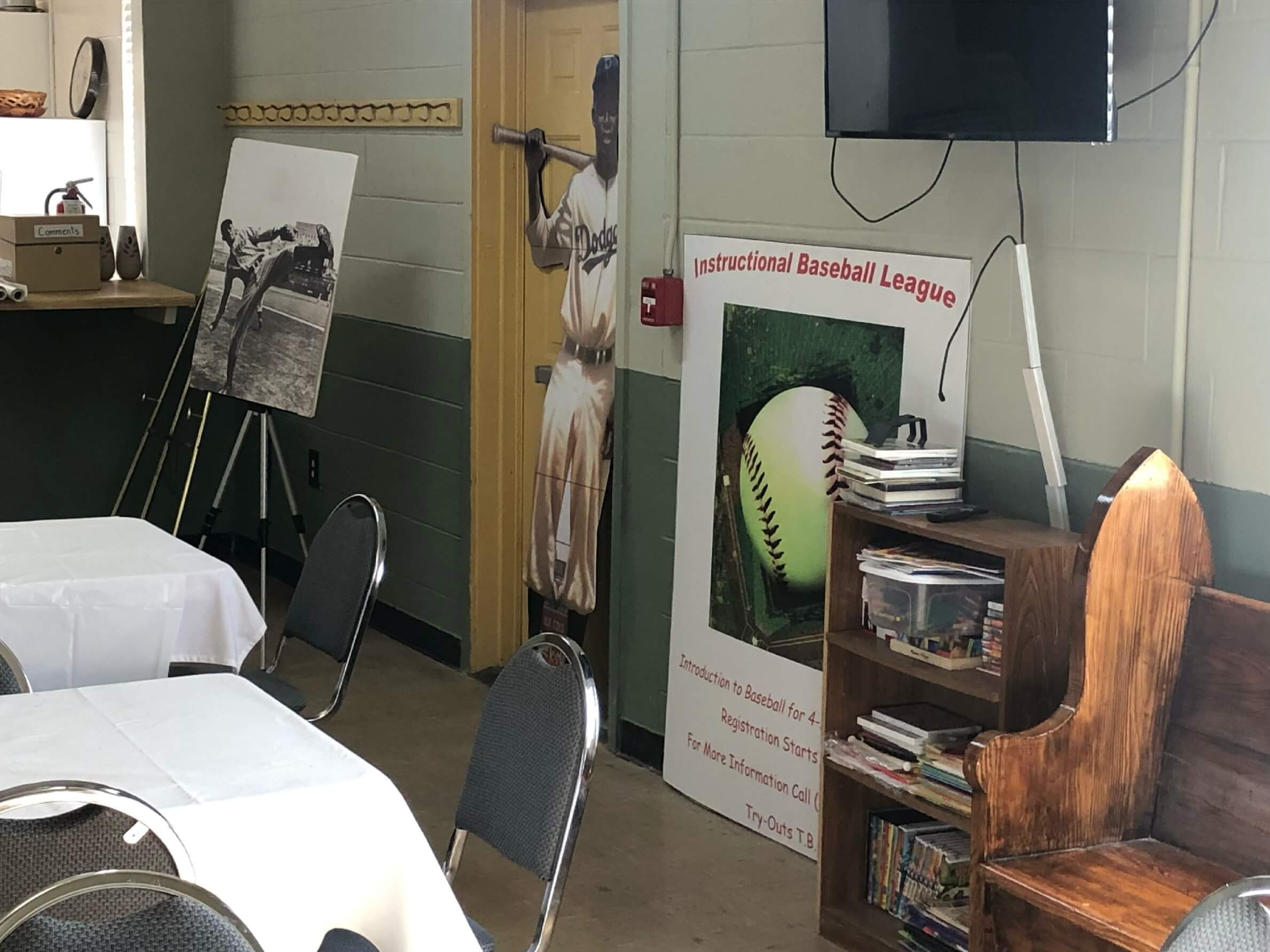 An indoor area featuring a life-sized Jackie Robinson cutout, a bookshelf, and a large poster for an "Instructional Baseball League."
