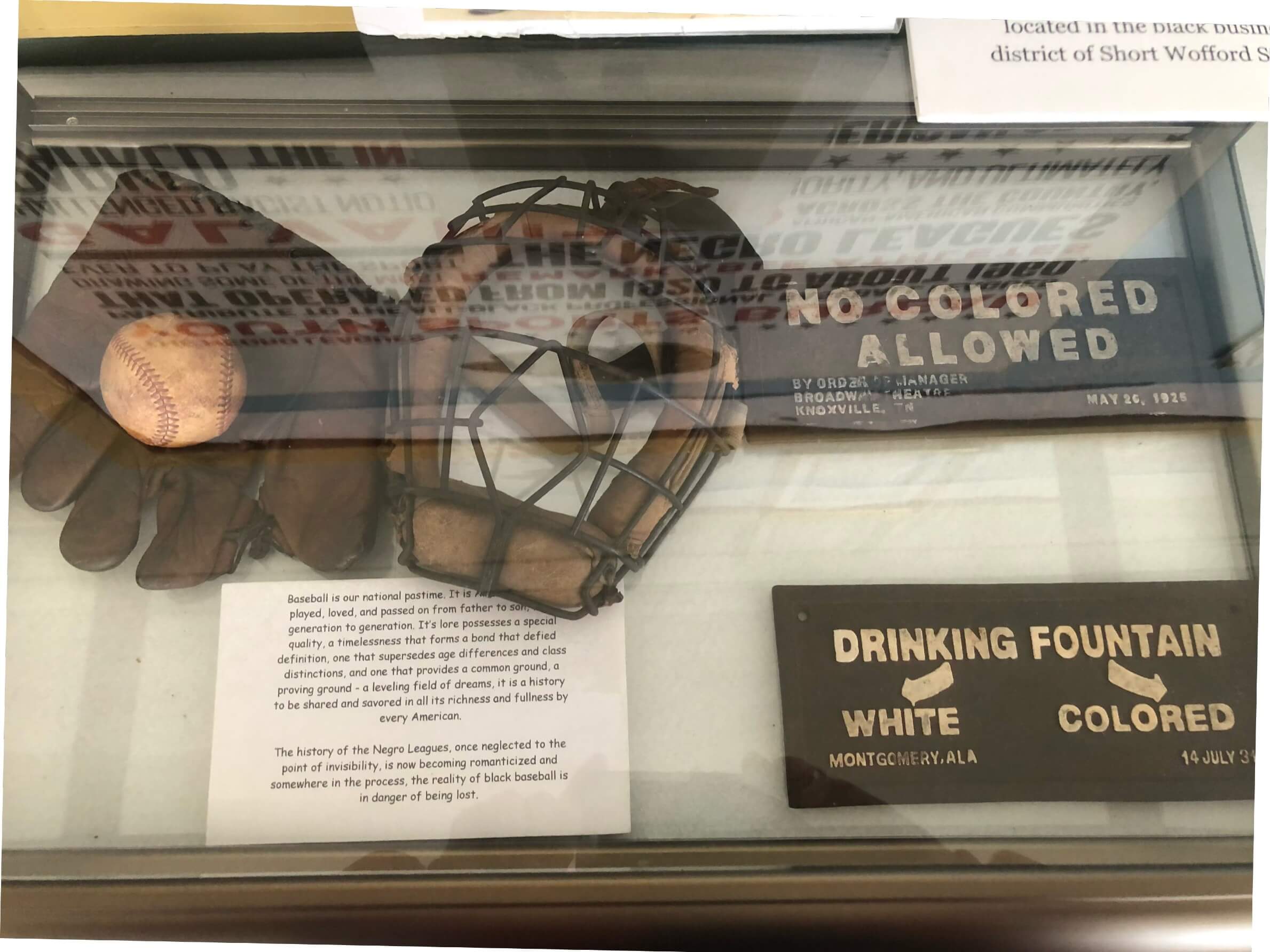 A glass display case containing an antique catcher's mask, glove, and baseball, alongside historical "No Colored Allowed" and "White/Colored" drinking fountain signs.
